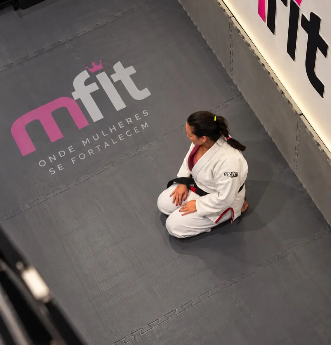Vista de cima do tatame da MFIT com a frase onde mulheres se fortalecem e uma aluna de kimono sentada