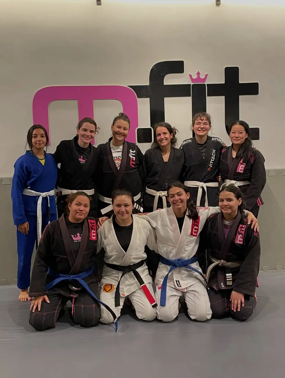 Alunas do jiu-jitsu feminino da MFIT de kimono em frente ao logo da academia
