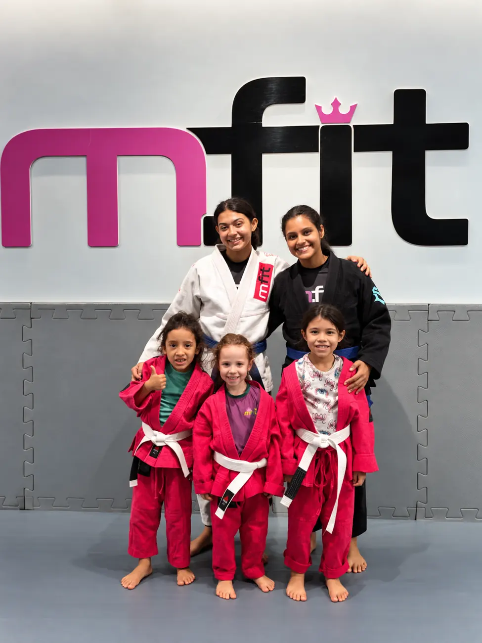 Três meninas de kimono rosa com duas professoras em frente ao logo da MFIT