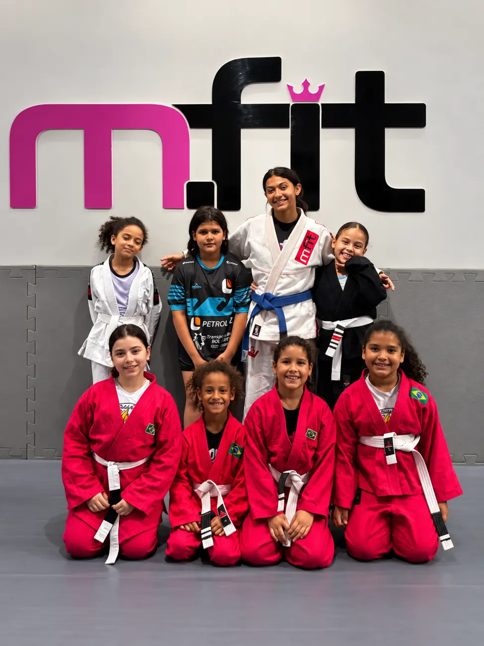 Turma de meninas do MFIT Kids de kimono em frente ao logo da MFIT